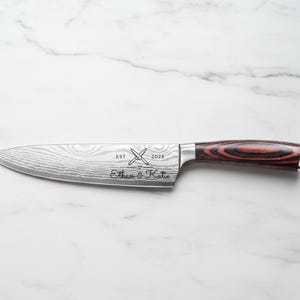 Personalized chef knife with engraved names "Ethan & Katie," crossed knife design, and "EST 2023" on a patterned stainless steel blade, featuring a red and black woodgrain handle on a white marble background.