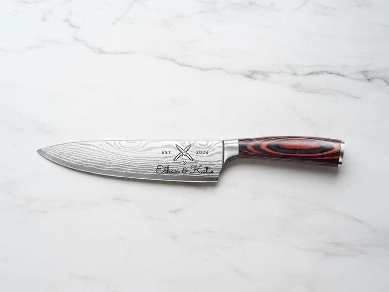 Personalized chef knife with engraved names "Ethan & Katie," crossed knife design, and "EST 2023" on a patterned stainless steel blade, featuring a red and black woodgrain handle on a white marble background.