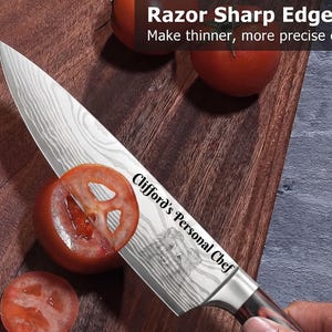 Personalized Damascus-style chef knife slicing a tomato on a wooden cutting board, custom laser engraved with the text "Clifford's Personal Chef" and a detailed dog portrait on the blade. Fresh tomatoes and a hand holding the knife demonstrate its razor-sharp edge in a kitchen setting.