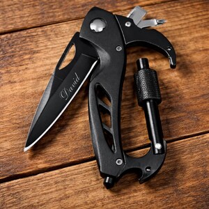 A black multi-tool knife with a carabiner is shown lying flat on a wooden surface. The blade is partially open and features the word "David" engraved on it. The handle is ergonomic and has several functions, including a Phillips-head screwdriver, a flathead screwdriver, and a hexagonal wrench. The multi-tool is positioned diagonally, with the carabiner on the right and the knife blade on the left. The lighting is bright and even.