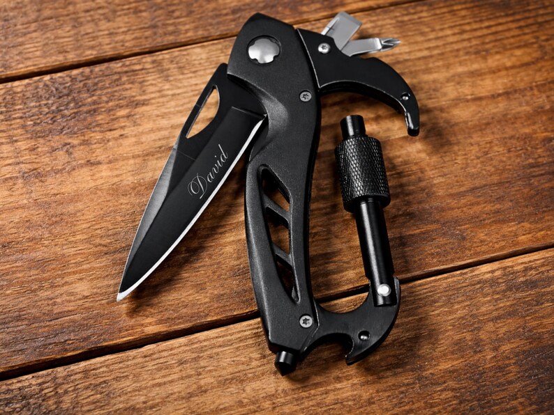 A black multi-tool knife with a carabiner is shown lying flat on a wooden surface. The blade is partially open and features the word "David" engraved on it. The handle is ergonomic and has several functions, including a Phillips-head screwdriver, a flathead screwdriver, and a hexagonal wrench. The multi-tool is positioned diagonally, with the carabiner on the right and the knife blade on the left. The lighting is bright and even.