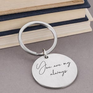 A silver keychain with a round pendant. The pendant has the inscription "You are my always" in a cursive font. The keychain is in front of a stack of books.