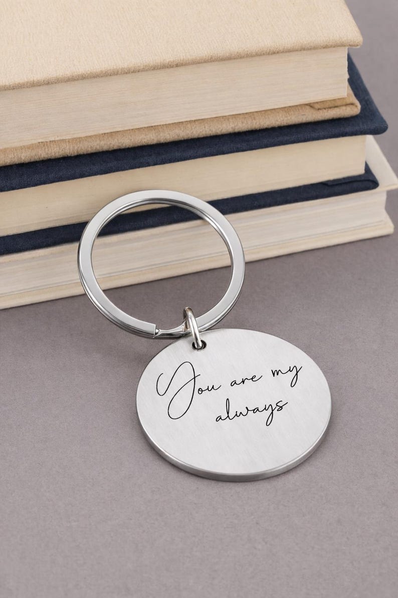 A silver keychain with a round pendant. The pendant has the inscription "You are my always" in a cursive font. The keychain is in front of a stack of books.