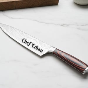 Custom engraved chef knife with the name “Chef Ethan” on a patterned stainless steel blade, featuring a red and black woodgrain handle, displayed on a white marble countertop with a wooden cutting board in the background.