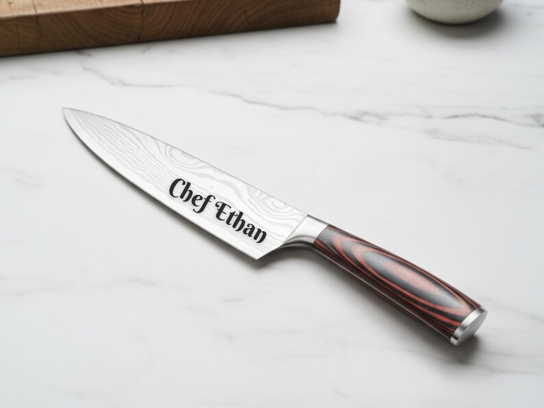 Custom engraved chef knife with the name “Chef Ethan” on a patterned stainless steel blade, featuring a red and black woodgrain handle, displayed on a white marble countertop with a wooden cutting board in the background.
