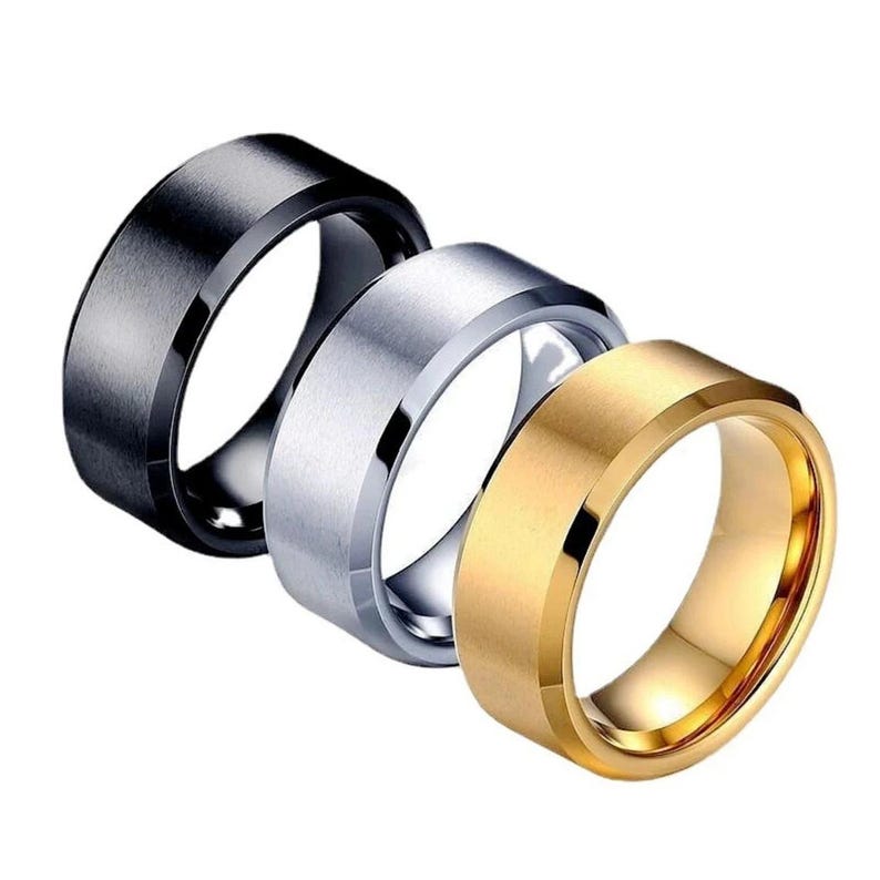 Three rings in a row, each with a different color. The first ring is silver, the second is black, and the third is gold. All three rings have a flat, wide band design with a polished finish. The rings are likely made of metal.