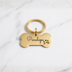 Personalized gold dog tag with engraved name Penelope in cursive. Custom bone-shaped brass pet ID tag with paw print cutout and gold keyring on a marble surface.