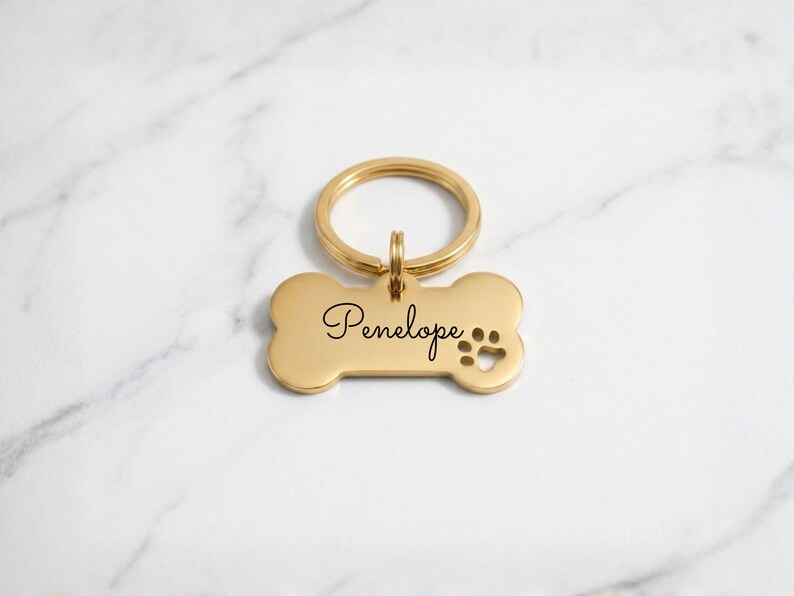 Personalized gold dog tag with engraved name Penelope in cursive. Custom bone-shaped brass pet ID tag with paw print cutout and gold keyring on a marble surface.
