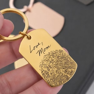 Close-up of a personalized gold fingerprint keychain held between fingers. The dog-tag-style keychain features the engraved handwritten message “Love, Mom” above a detailed fingerprint engraving. The polished gold finish and custom design are highlighted against a softly blurred background, showcasing a meaningful keepsake gift for memorials, mothers, and loved ones.