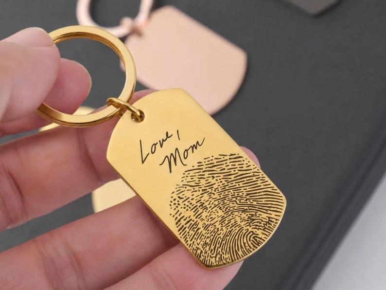 Close-up of a personalized gold fingerprint keychain held between fingers. The dog-tag-style keychain features the engraved handwritten message “Love, Mom” above a detailed fingerprint engraving. The polished gold finish and custom design are highlighted against a softly blurred background, showcasing a meaningful keepsake gift for memorials, mothers, and loved ones.