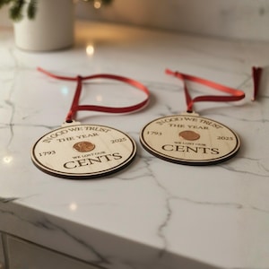 May include: Two wooden Christmas ornaments with a penny in the center. The ornaments have a red ribbon for hanging and feature the text "IN GOD WE TRUST THE YEAR" and "WE LOST OUR CENTS" with the years 1793 and 2025.