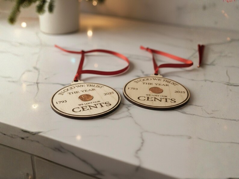 May include: Two wooden Christmas ornaments with a penny in the center. The ornaments have a red ribbon for hanging and feature the text "IN GOD WE TRUST THE YEAR" and "WE LOST OUR CENTS" with the years 1793 and 2025.