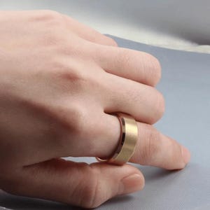A gold-colored ring with a flat top and beveled edges. The ring is worn on a finger, showcasing its simple, modern design. The ring's surface has a matte finish, reflecting light subtly.
