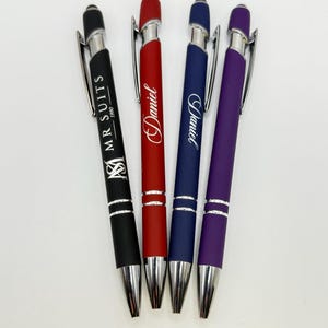 A set of four customized metal ballpoint pens in different colors, lying on a white background. From left to right, the pens are: Black (engraved with a logo and "MR SUITS 1980"), Red (engraved with the name "Daniel" in script), Blue (engraved with the name "David" in script), and Purple (unengraved). All pens have silver metal clips, tips, and mid-sections.