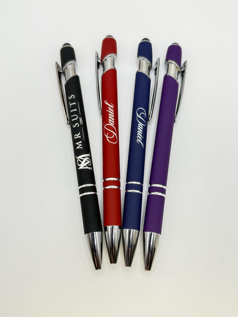 A set of four customized metal ballpoint pens in different colors, lying on a white background. From left to right, the pens are: Black (engraved with a logo and "MR SUITS 1980"), Red (engraved with the name "Daniel" in script), Blue (engraved with the name "David" in script), and Purple (unengraved). All pens have silver metal clips, tips, and mid-sections.