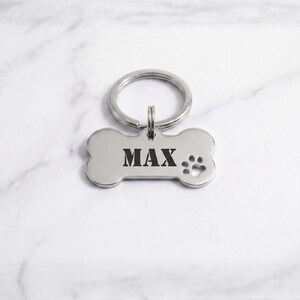 Custom silver bone-shaped pet ID tag engraved with the name 'MAX' in a bold serif stencil font, featuring a matching silver split key ring and paw print cutout on a white marble background.