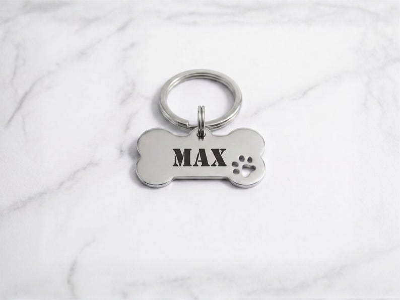 Custom silver bone-shaped pet ID tag engraved with the name 'MAX' in a bold serif stencil font, featuring a matching silver split key ring and paw print cutout on a white marble background.