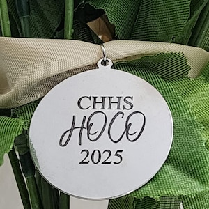 Puede incluir: Un colgante redondo plateado con el texto "CHHS HOCO 2025" en escritura negra. El colgante cuelga de una cinta verde y beige, rodeado de follaje verde.