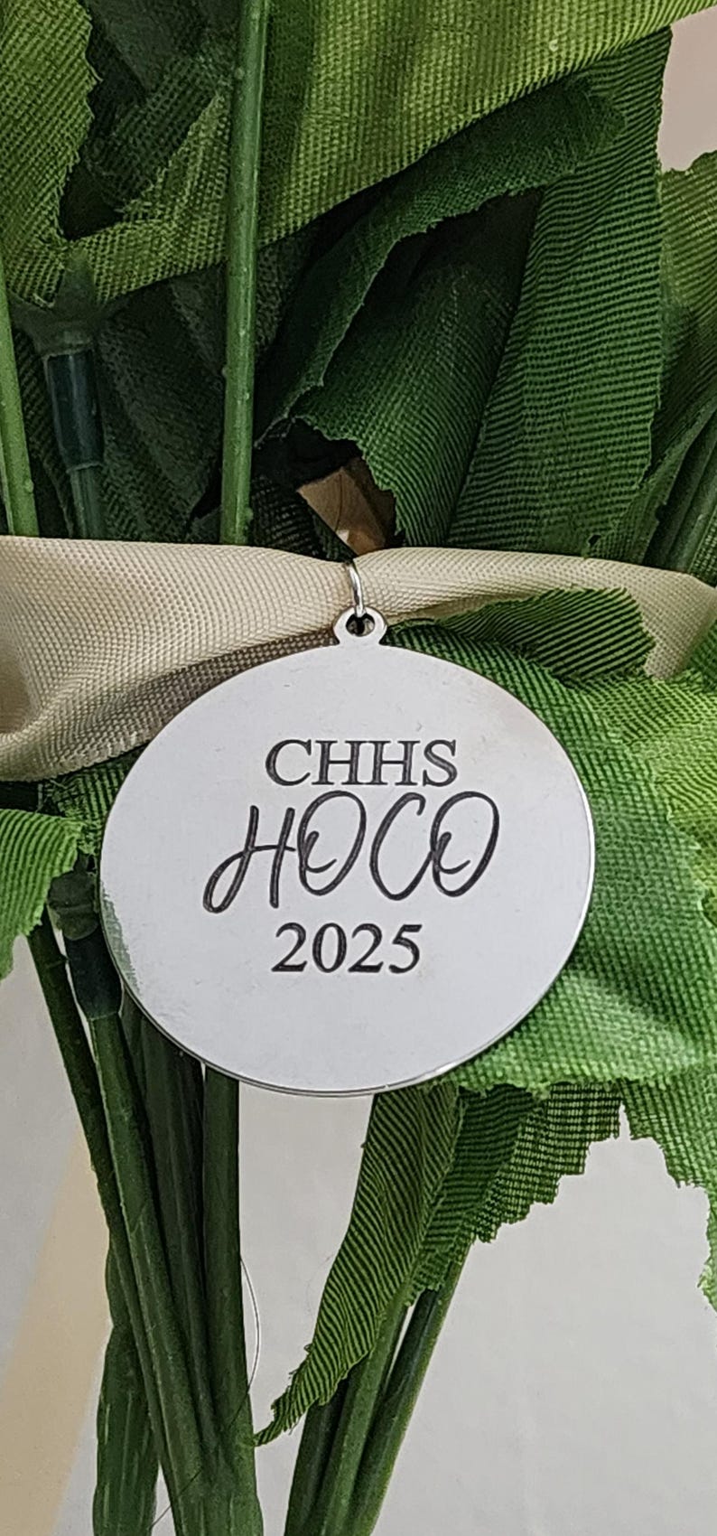 A high-resolution close-up of a circular silver metal charm attached to a bouquet of green leaves and a tan ribbon. The charm is permanently laser-engraved with "CHHS" in a bold serif font, "HOCO" in an elegant cursive script, and "2025" at the bottom. The metal has a polished, reflective finish, showcasing the precision of the dark engraving.