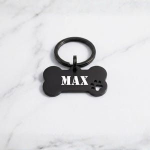 Custom black bone-shaped pet ID tag engraved with the name 'MAX' in white stencil font, featuring a matching black split key ring and paw print cutout on a white marble surface.