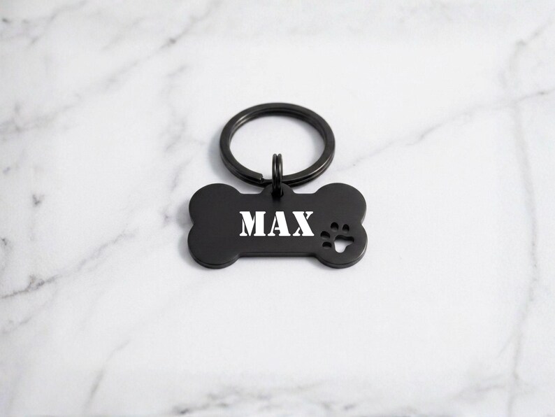 Custom black bone-shaped pet ID tag engraved with the name 'MAX' in white stencil font, featuring a matching black split key ring and paw print cutout on a white marble surface.