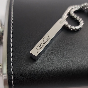 A close-up, high-angle shot of a rectangular silver pendant engraved with the name "Michael" in a script font, resting on a black, leather-wrapped hip flask. The pendant is attached to a silver chain, which is partially visible on the right. The black flask has white stitching visible along its edge.