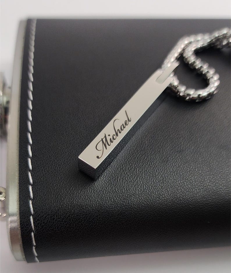 A close-up, high-angle shot of a rectangular silver pendant engraved with the name "Michael" in a script font, resting on a black, leather-wrapped hip flask. The pendant is attached to a silver chain, which is partially visible on the right. The black flask has white stitching visible along its edge.
