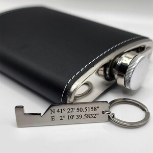 Black leather-wrapped flask with a stainless steel cap and a silver-colored keychain. The keychain has the coordinates "N 41° 22' 50.5158" E 2° 10' 39.5832" engraved on it. White stitching accents the flask.