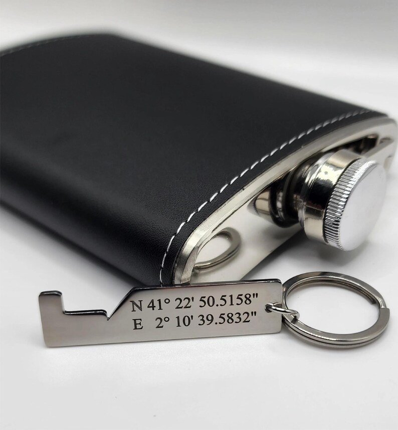 Black leather-wrapped flask with a stainless steel cap and a silver-colored keychain. The keychain has the coordinates "N 41° 22' 50.5158" E 2° 10' 39.5832" engraved on it. White stitching accents the flask.