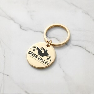 May include: A gold-colored keychain with a round pendant. The pendant features the text "GREEN VALLEY PREMIUM PROPERTIES" and a black silhouette of a house. The keychain is on a white marble surface.