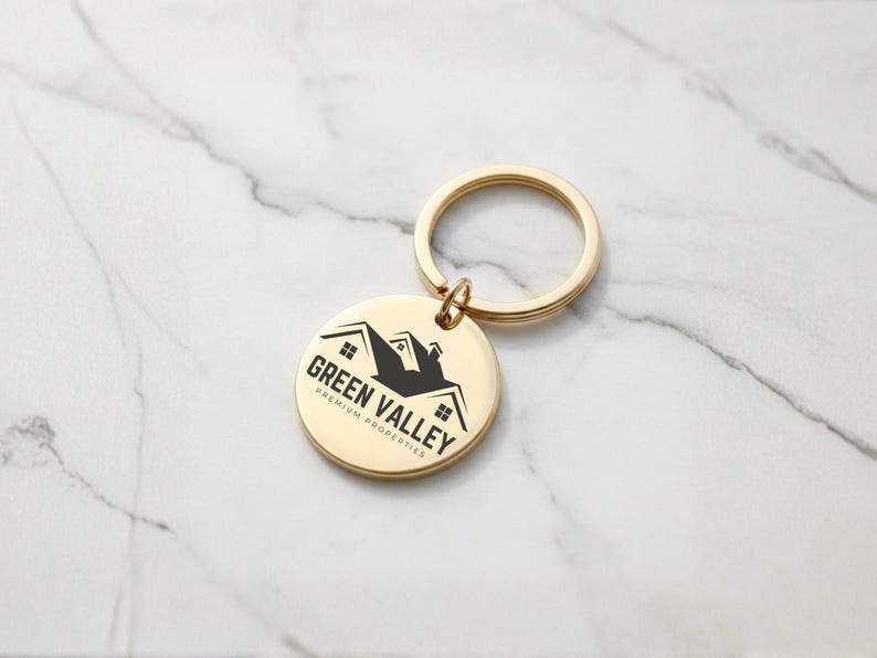 May include: A gold-colored keychain with a round pendant. The pendant features the text "GREEN VALLEY PREMIUM PROPERTIES" and a black silhouette of a house. The keychain is on a white marble surface.