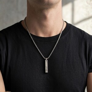K&ouml;nnte beinhalten: Halskette mit silberfarbenem Anh&auml;nger. Der rechteckige Anh&auml;nger h&auml;ngt an einer feinen Kette. Die Kette wird &uuml;ber einem schwarzen T-Shirt getragen. Der Anh&auml;nger hat eine Inschrift. Ein minimalistisches und stilvolles Schmuckst&uuml;ck.