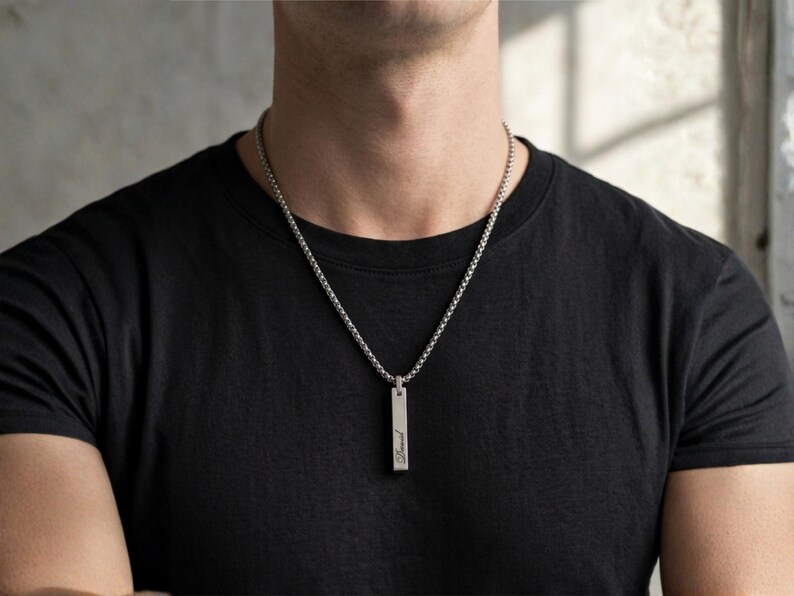 K&ouml;nnte beinhalten: Halskette mit silberfarbenem Anh&auml;nger. Der rechteckige Anh&auml;nger h&auml;ngt an einer feinen Kette. Die Kette wird &uuml;ber einem schwarzen T-Shirt getragen. Der Anh&auml;nger hat eine Inschrift. Ein minimalistisches und stilvolles Schmuckst&uuml;ck.