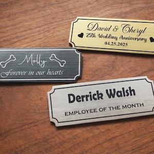Three engraved metal nameplates displayed on a wooden table: a black pet memorial plaque reading “Molly – Forever in our hearts” with bone icons, a gold “David & Cheryl – 25th Wedding Anniversary – 04.25.2025” plate with heart symbols, and a silver “Derrick Walsh – Employee of the Month” nameplate, all with decorative borders.