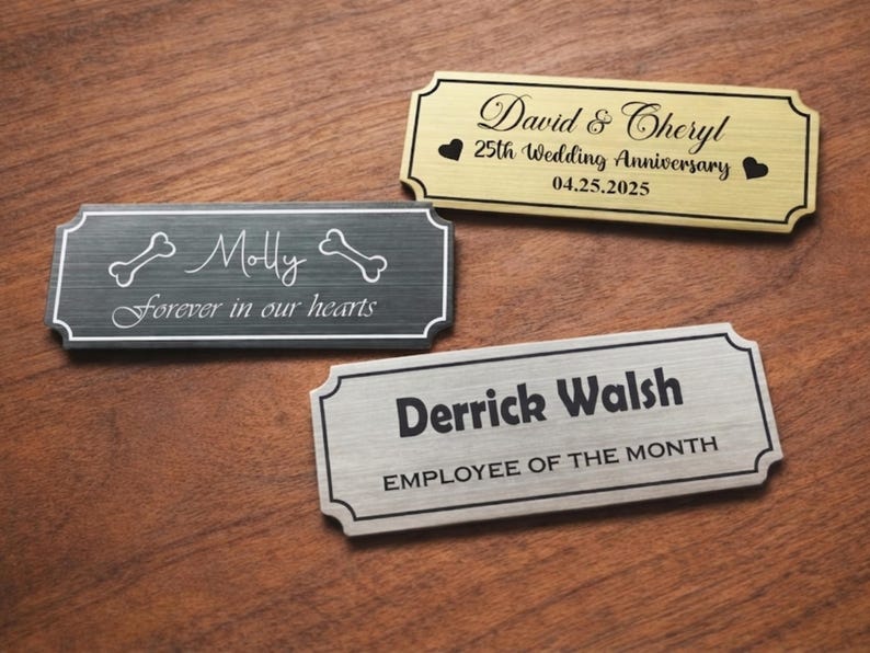 Three engraved metal nameplates displayed on a wooden table: a black pet memorial plaque reading “Molly – Forever in our hearts” with bone icons, a gold “David & Cheryl – 25th Wedding Anniversary – 04.25.2025” plate with heart symbols, and a silver “Derrick Walsh – Employee of the Month” nameplate, all with decorative borders.