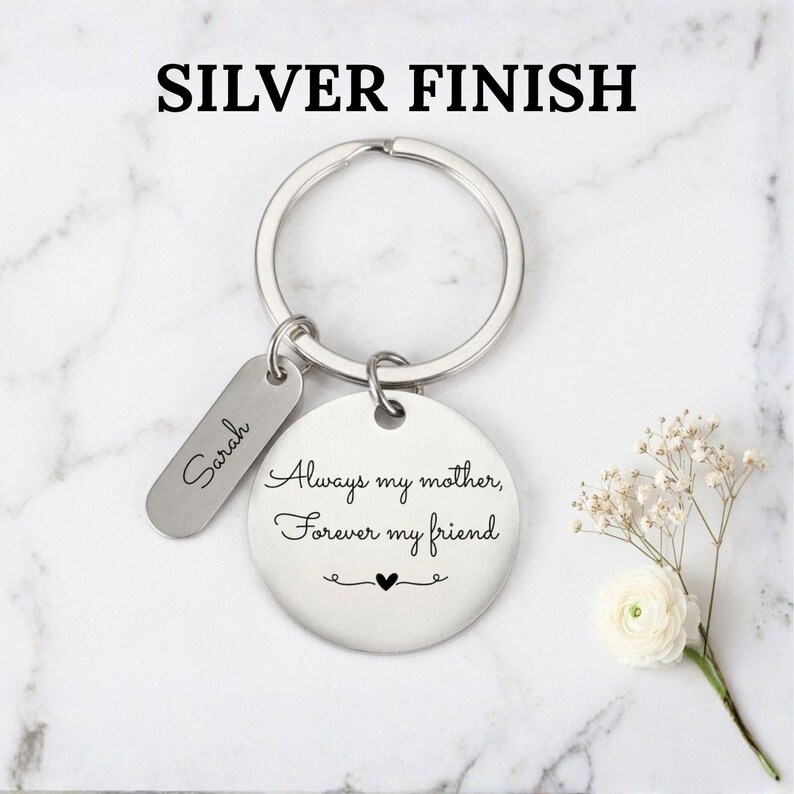 Silver finish personalized keychain for mom with engraved message “Always my mother, forever my friend” and custom name tag, displayed on a marble background with delicate white flowers.