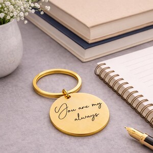 Gold round engraved keychain displayed on a desk beside a spiral notebook, fountain pen, stacked books, and a vase of white flowers. The keychain features the handwritten message “You are my always” engraved on a brushed gold metal pendant. Soft natural lighting and elegant desk décor create a warm, sentimental gift presentation suitable for anniversaries, weddings, or loved ones.