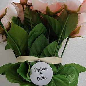K&ouml;nnte beinhalten: Ein Strau&szlig; aus k&uuml;nstlichen blassrosa Rosen mit gr&uuml;nen Bl&auml;ttern, zusammengebunden mit einem hellbeigen Band. Ein kreisf&ouml;rmiges silberfarbenes Metallschild h&auml;ngt am Band und zeigt den schwarzen Aufdruck Stephanie & Colton in Schreibschrift.