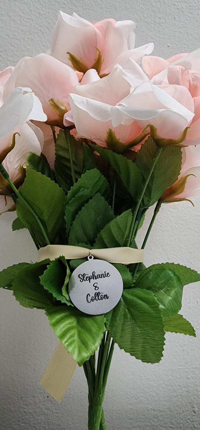 K&ouml;nnte beinhalten: Ein Strau&szlig; aus k&uuml;nstlichen blassrosa Rosen mit gr&uuml;nen Bl&auml;ttern, zusammengebunden mit einem hellbeigen Band. Ein kreisf&ouml;rmiges silberfarbenes Metallschild h&auml;ngt am Band und zeigt den schwarzen Aufdruck Stephanie & Colton in Schreibschrift.