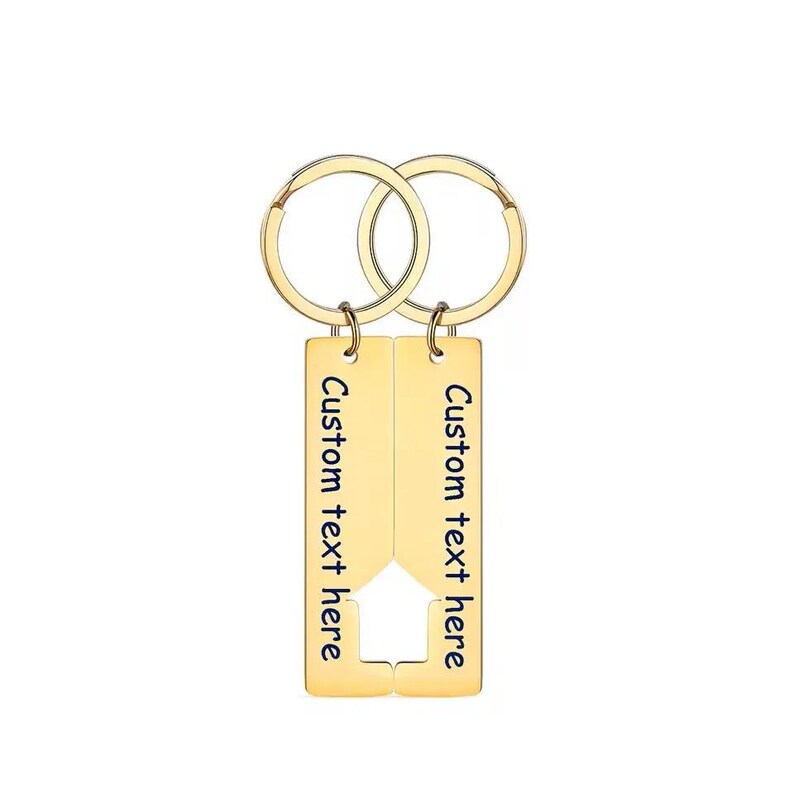 Two gold-colored keychains with a house-shaped design. Each keychain has a rectangular tag with the text "Custom text here" in blue. The keychains are connected by two gold-colored key rings.