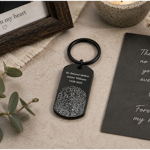 Memorial fingerprint keychain displayed in a styled remembrance setting. The black dog-tag-style keychain is engraved with “My Beloved Mother Ashley Williams 1938–2025” above a detailed fingerprint design. It rests on a neutral stone surface beside eucalyptus leaves, a candle, a framed photo with the words “Forever in my heart,” and a sympathy card, creating a warm and heartfelt memorial tribute scene.