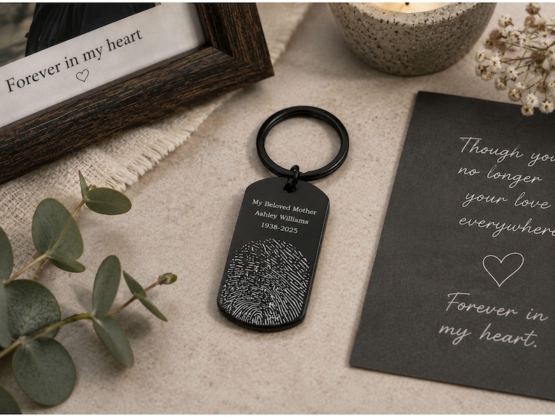 Memorial fingerprint keychain displayed in a styled remembrance setting. The black dog-tag-style keychain is engraved with “My Beloved Mother Ashley Williams 1938–2025” above a detailed fingerprint design. It rests on a neutral stone surface beside eucalyptus leaves, a candle, a framed photo with the words “Forever in my heart,” and a sympathy card, creating a warm and heartfelt memorial tribute scene.