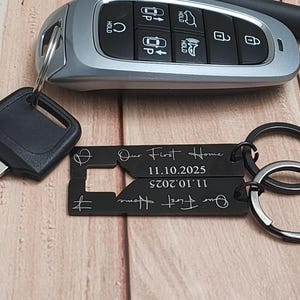 Black metal keychains with the text "Our First Home" and the date 11.10.2025. The keychains are attached to a silver key ring. A car key is in the background.