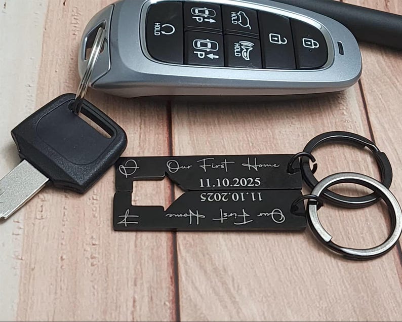 Black metal keychains with the text "Our First Home" and the date 11.10.2025. The keychains are attached to a silver key ring. A car key is in the background.