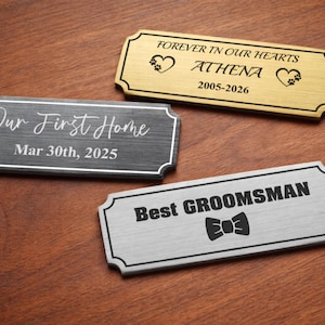 Three engraved metal nameplates on a wooden surface: a gold pet memorial plaque reading “Forever in our hearts – Athena – 2005–2026” with paw print hearts, a black “Our First Home – Mar 30th, 2025” plaque in script font, and a silver “Best Groomsman” plate with a bowtie icon, all with decorative borders.