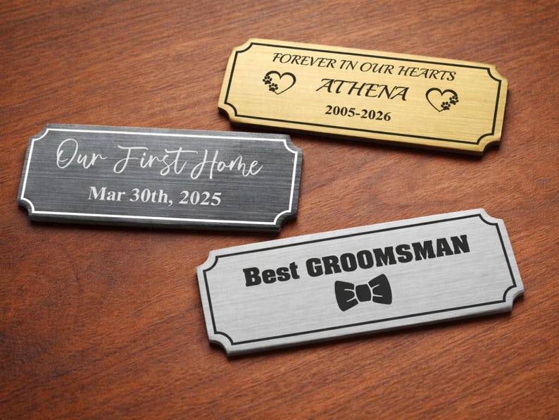 Three engraved metal nameplates on a wooden surface: a gold pet memorial plaque reading “Forever in our hearts – Athena – 2005–2026” with paw print hearts, a black “Our First Home – Mar 30th, 2025” plaque in script font, and a silver “Best Groomsman” plate with a bowtie icon, all with decorative borders.