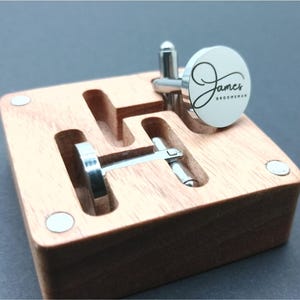 Puede incluir: Gemelos plateados con una cara circular blanca, grabados con el nombre "James" y la palabra "Groomman" en escritura negra. Los gemelos se muestran en una caja de madera con huecos para cada gemelo.
