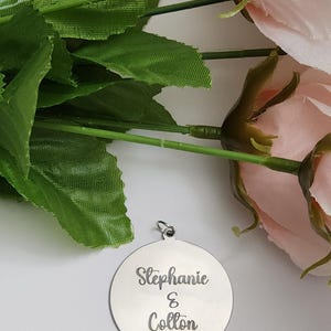 K&ouml;nnte beinhalten: Ein runder, silberfarbener Metallanh&auml;nger mit einem kleinen Verbindungsring an der Oberseite. Die Oberfl&auml;che ist mit dem Text "Stephanie & Colton" in einer kursiven Schriftart graviert. Der Anh&auml;nger liegt neben einem Strau&szlig; k&uuml;nstlicher rosa Rosen.