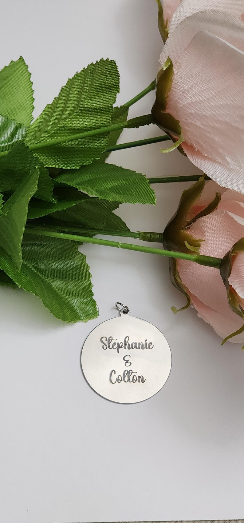 K&ouml;nnte beinhalten: Ein runder, silberfarbener Metallanh&auml;nger mit einem kleinen Verbindungsring an der Oberseite. Die Oberfl&auml;che ist mit dem Text "Stephanie & Colton" in einer kursiven Schriftart graviert. Der Anh&auml;nger liegt neben einem Strau&szlig; k&uuml;nstlicher rosa Rosen.