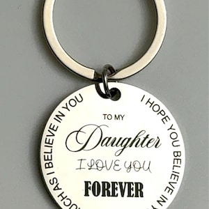 Close-up of a round silver keychain with a white engraved pendant. The pendant reads “To my Daughter I love you forever” in mixed script and bold fonts, with a circular border text that says, “I hope you believe in yourself as much as I believe in you.”